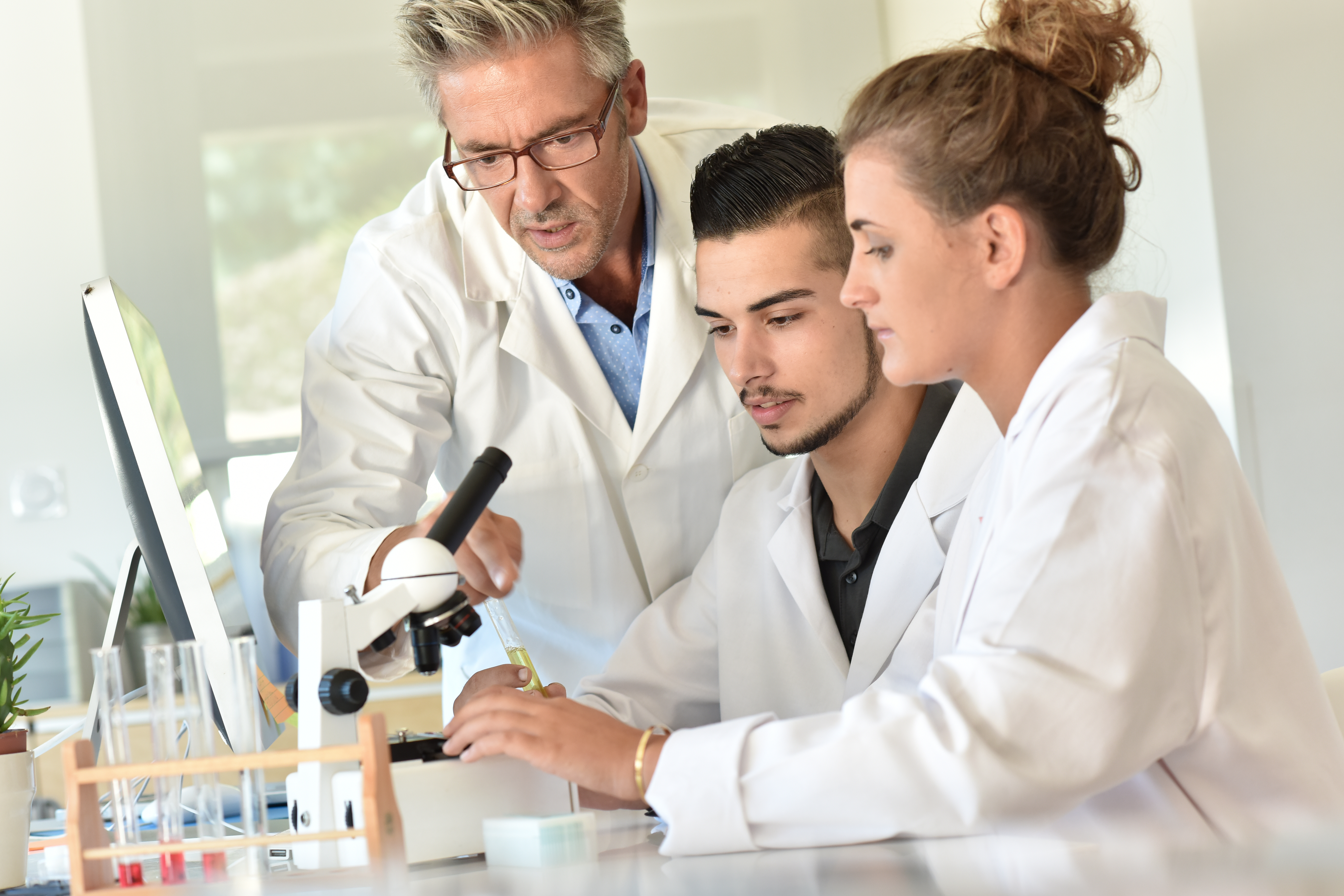 Teacher in a lab coat helping students in lab coats to use a microscope
