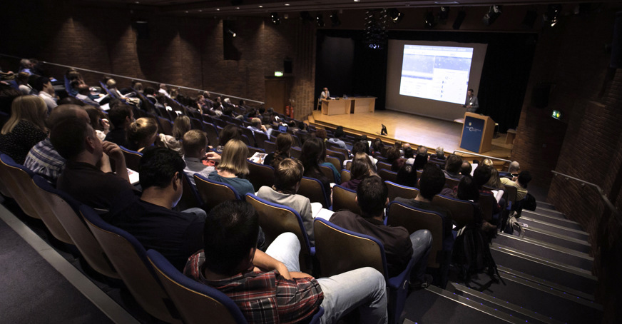 View from the back of lecture theater with students in the seats and a lecturer on stage in front of a screen